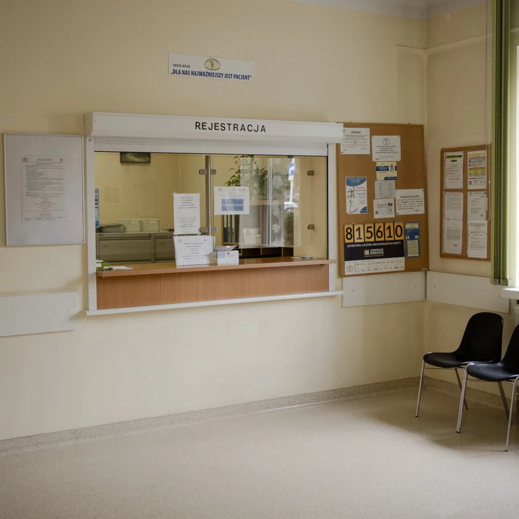 Wnętrze przychodni – okienko rejestracji z napisem 'Rejestracja', tablica ogłoszeń z informacjami dla pacjentów oraz dwa krzesła w poczekalni