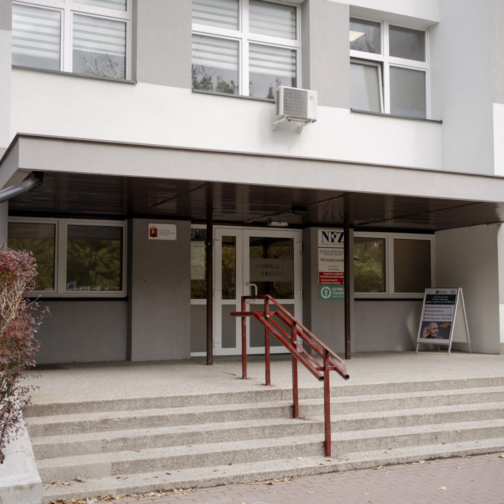 Wejście do przychodni lekarskiej z szyldem NFZ i czerwonymi poręczami przy schodach