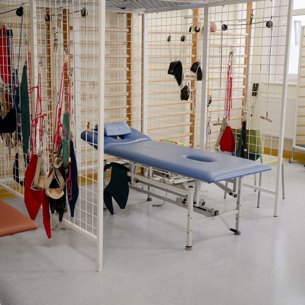 Sala rehabilitacyjna z niebieskim stołem do ćwiczeń i zestawem linek oraz pasów terapeutycznych